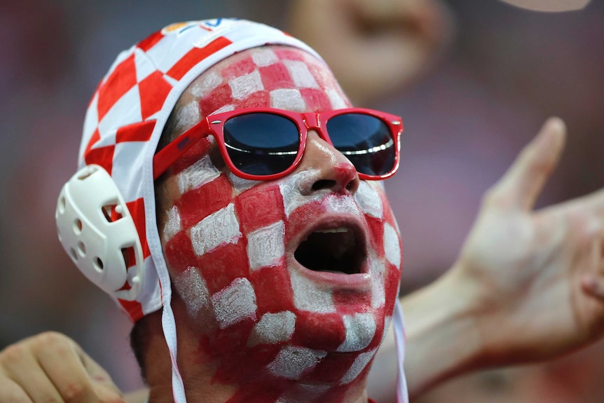 A Croatian fan wearing red and white checked face paint, sun glasses and a water polo cap sings