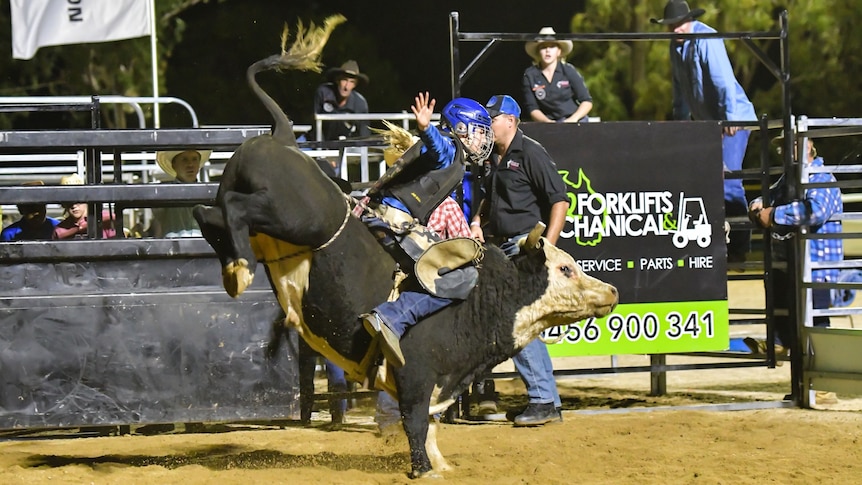 Bonnie on top of a black bucking bull wearing protective clothing and a helmet with one hand in the air