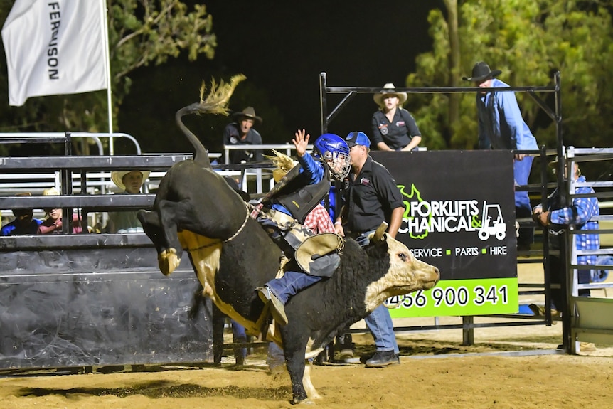 Bonnie on top of a black bucking bull wearing protective clothing and a helmet with one hand in the air