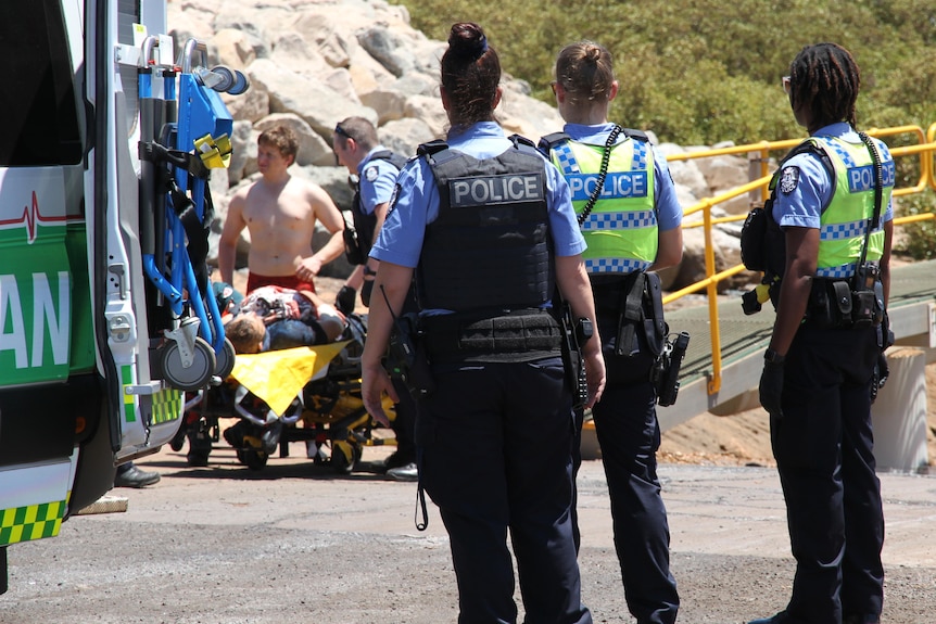 A group of police stand next to an ambulance, watching a man on a stretcher being attended to.