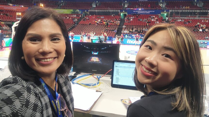 Patricia takes a selfie of her and Beatrice sitting at a commentating desk in a basketball stadium.