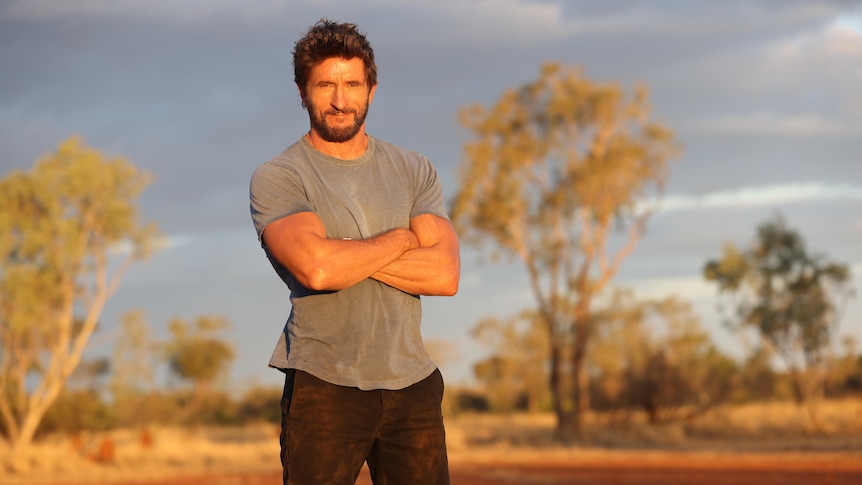 Australian Survivor host Jonathan LaPaglia stands with his arms crossed in the outback.