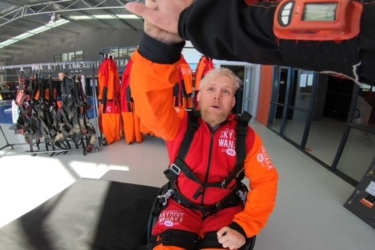 A man in a wheelchair wearing an orange skydive jumping suit hi-fiving someone who is behind the camera