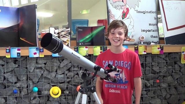 A boy stands beside a telescope in a classroom