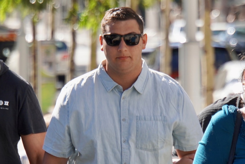 A mid-shot of a young man wearing a collared shirt and sunglasses outside.