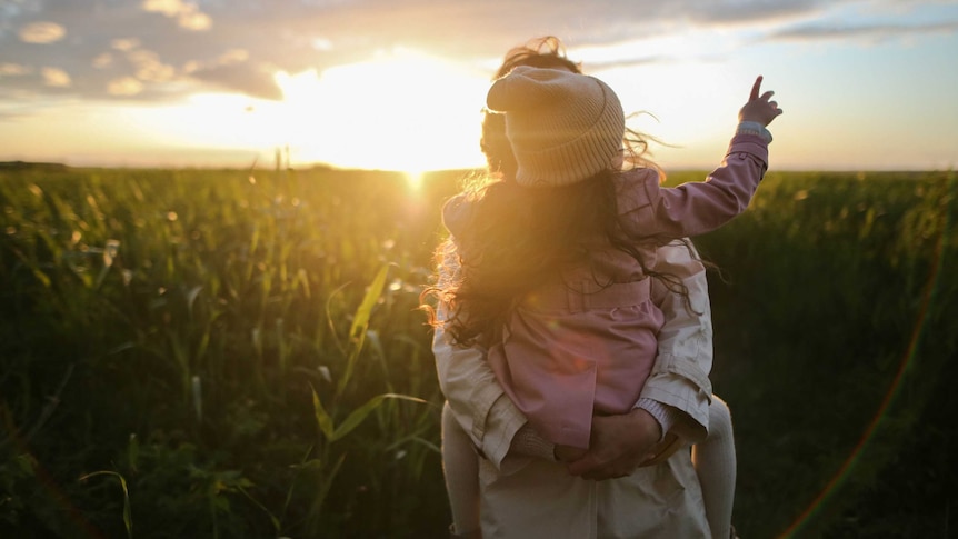 A woman holds a child in a field, surrounded by tall grass at dusk.