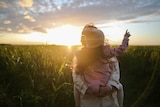 A woman holds a child in a field, surrounded by tall grass at dusk.