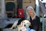 A smiling woman sits on the steps of her weatherboard home's verandah. A yellow labrador lays next to her, clutching a soft toy.