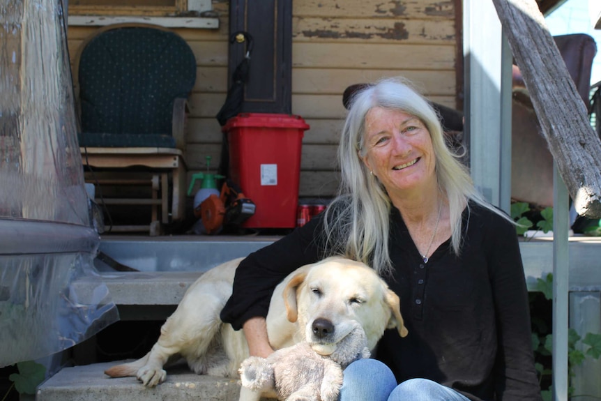 A smiling woman sits on the steps of her weatherboard home's verandah. A yellow labrador lays next to her, clutching a soft toy.