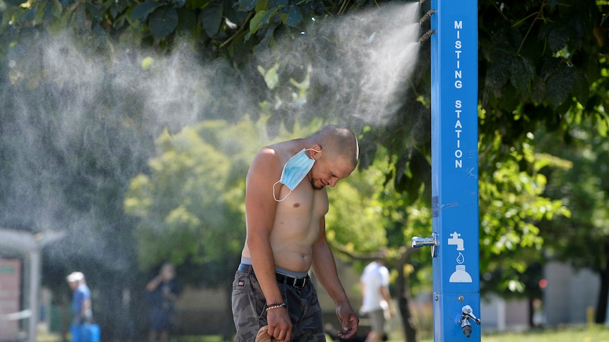 A shirtless man stands underneath a public misting fountain. 