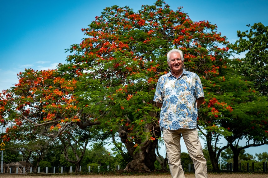 Man posing beside a tree with red flowers in a park on a sunny day.