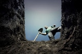 A man with a beard holding a shovel looks down into a freshly dug grave.