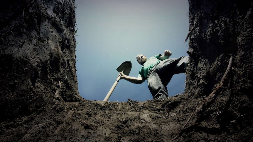 A man with a beard holding a shovel looks down into a freshly dug grave.