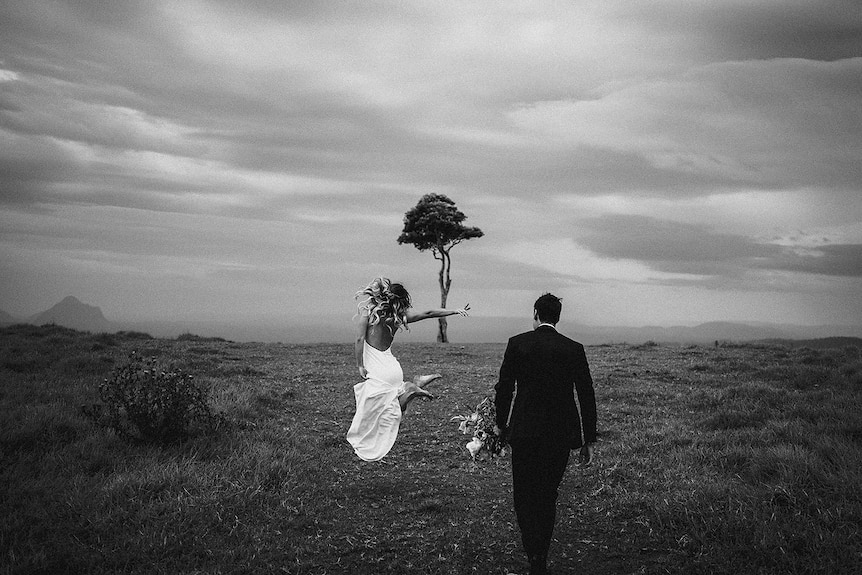 One Tree Hill black and white picture of bridge in dress jumping in the air while groom walks behind her