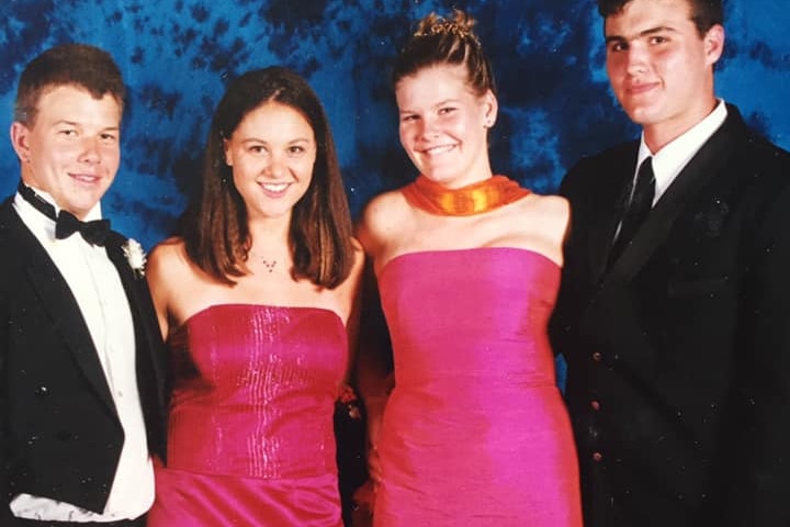 A photo of 4 teenagers, 2 wearing formal suits and 2 in formal dresses, against a dark blue marbled professional photo backdrop.