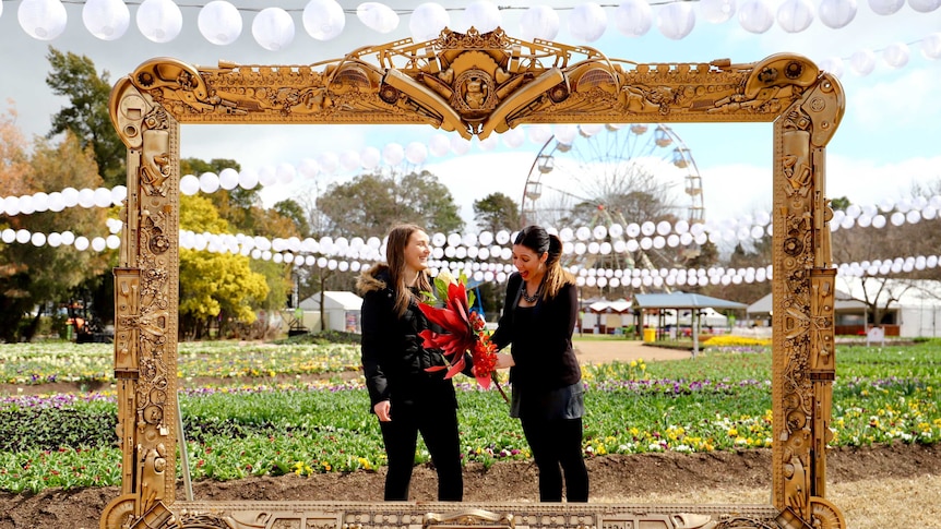 Two women standing in a frame with gardens of flowers and ferris wheel behind them