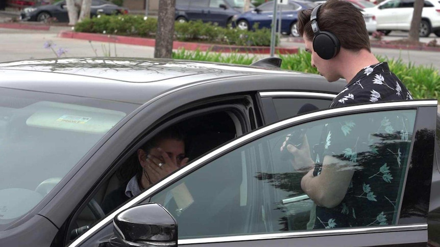 Ollie wearing headphones and holding a microphone confronts Lezlie in her car
