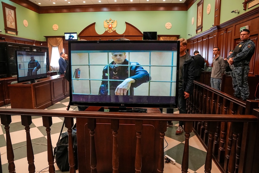 In a courtroom, an image of Alexei Navalny behind bars appears on a number of TV screens