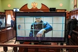 In a courtroom, an image of Alexei Navalny behind bars appears on a number of TV screens