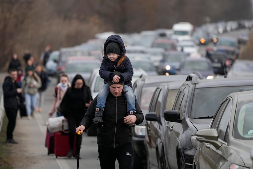 A child sits on a man's shoulders, as people pulling suitcases walk alongside a line of cars.