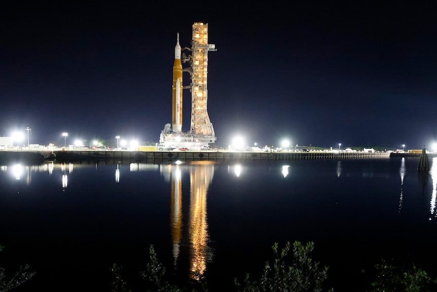 A rocket at a launch pad at night with lights reflecting on a lake in front