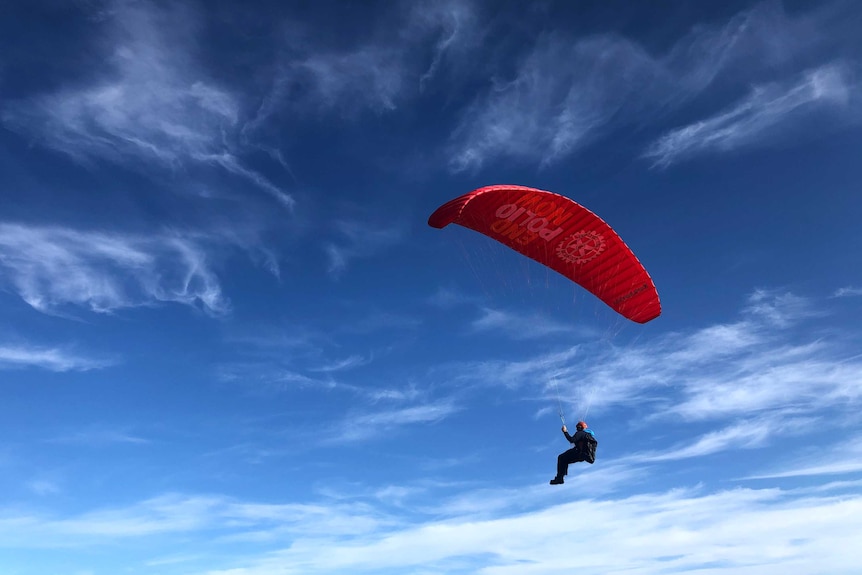A man paraglides with a red parachute with a dark blue sky with wispy clouds as his backdrop.