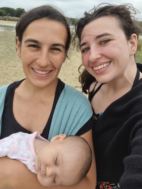 Two women smiling facing a camera on 'selfie' mode, with a beach in the background. One of the women is holding a sleeping baby.