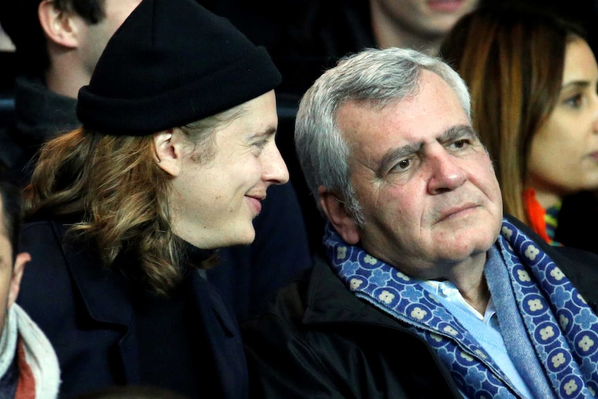 Thierry Herzog looks on while Pierre Sarkozy, with long hair and wearing a black beanie, smiles and speaks to him