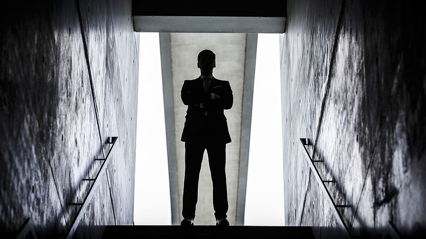 Man in business suit standing at the top of a staircase with arms folded with a shadow blocking out his face.