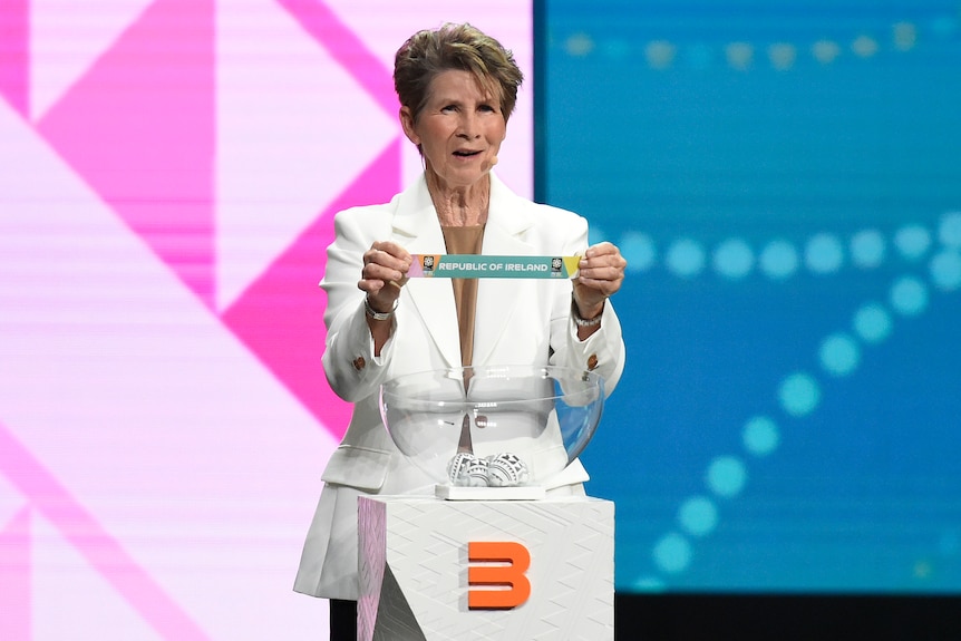 A woman in a white jacket holds up a piece of paper during a tournament draw