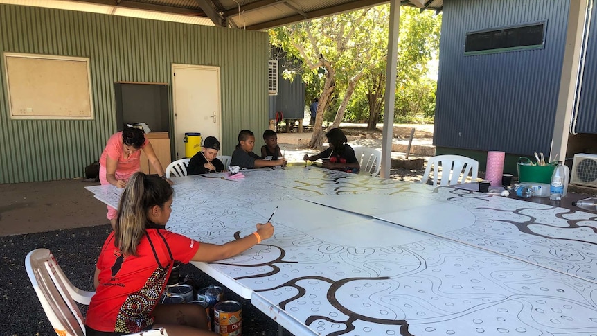 Children sit around a large unfinished mural painting in the outlines with colour
