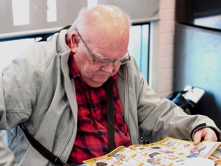 An elderly man sits at a table reading a catalogue. He is wearing a red and black plaid shirt and grey jacket.