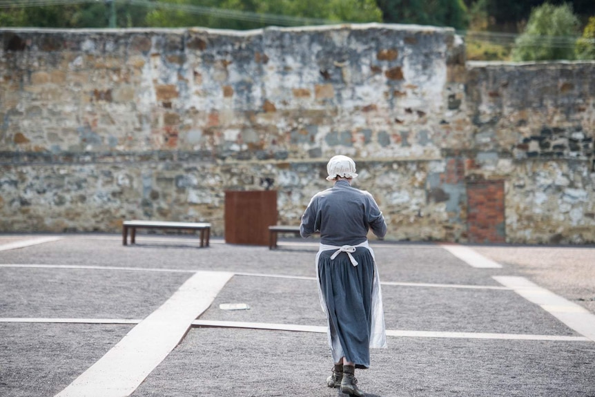 A woman in a bonnet and apron standing in a sandstone courtyard