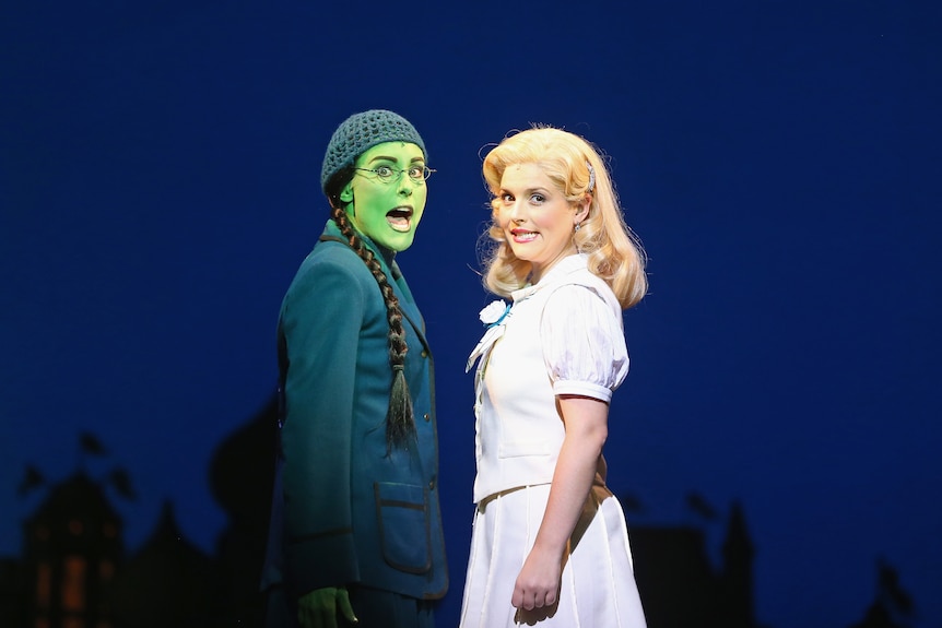 Sur scène, une femme en vert foncé à la peau peinte en vert chante aux côtés d'une femme blanche blonde portant un haut et une jupe blancs assortis.