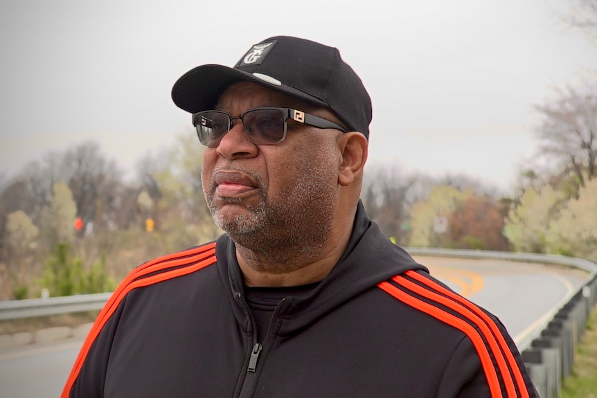 Derric wears a black baseball cap, sunglasses, and black hoodie with orange stripes as he stands by a road.