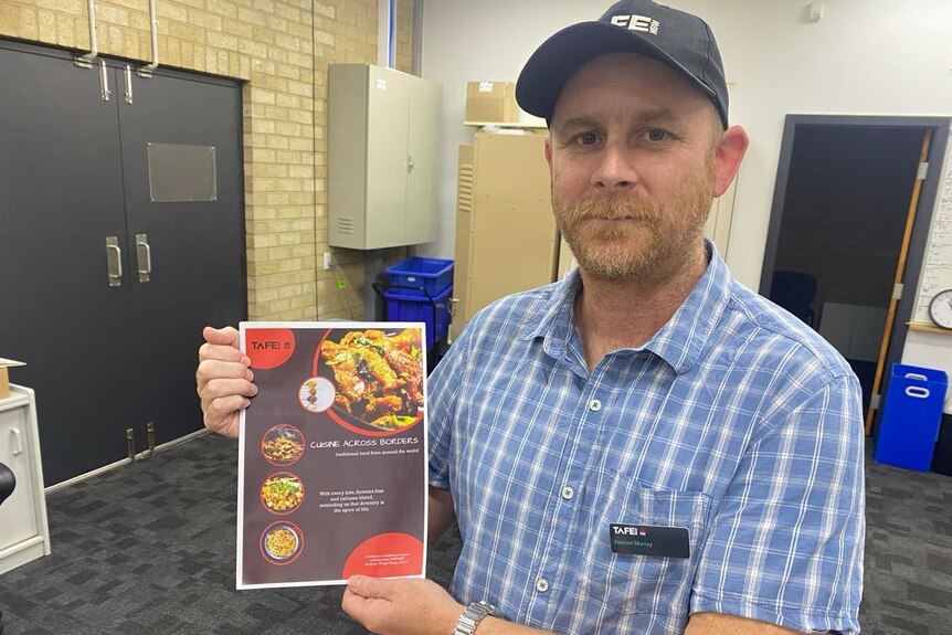 A man with a beard, a hat and short sleeves holds up a copy of a book called Cuisine Across Borders.
