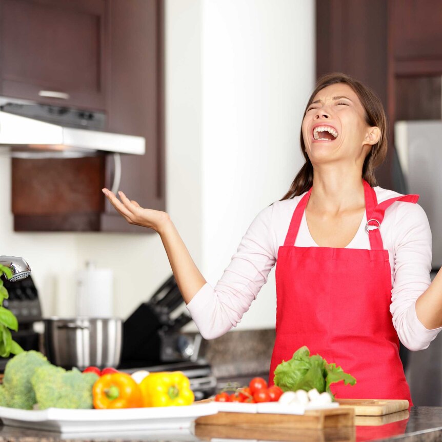 woman in red apron with hands in the air and look of frustration near kitchen bench laden with vegetables