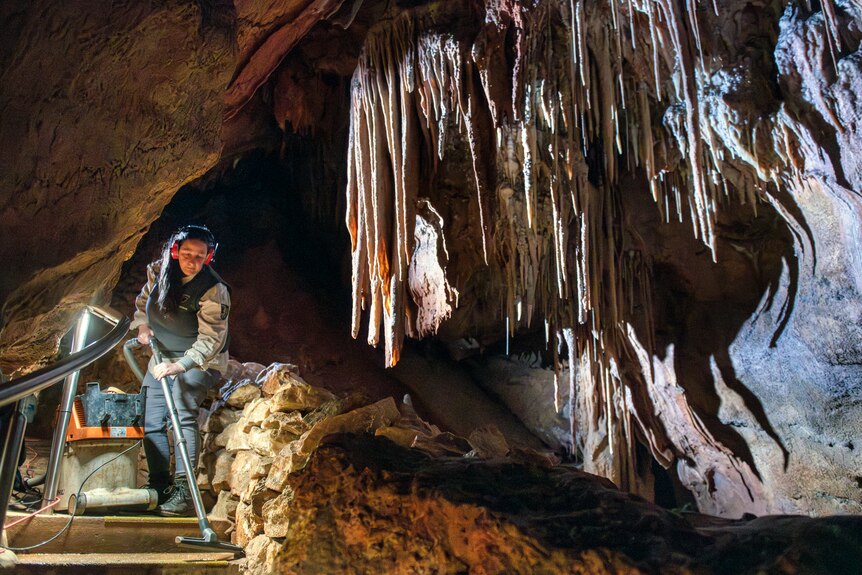 A woman wearing ear muffs vacuums the path alongside massive limestone and crystalline rock formations inside a cave.
