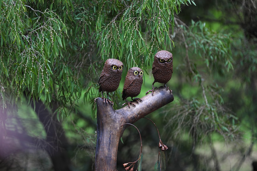 Metal sculpture of three owls sitting on a metal branch nestled in green tree leaves.