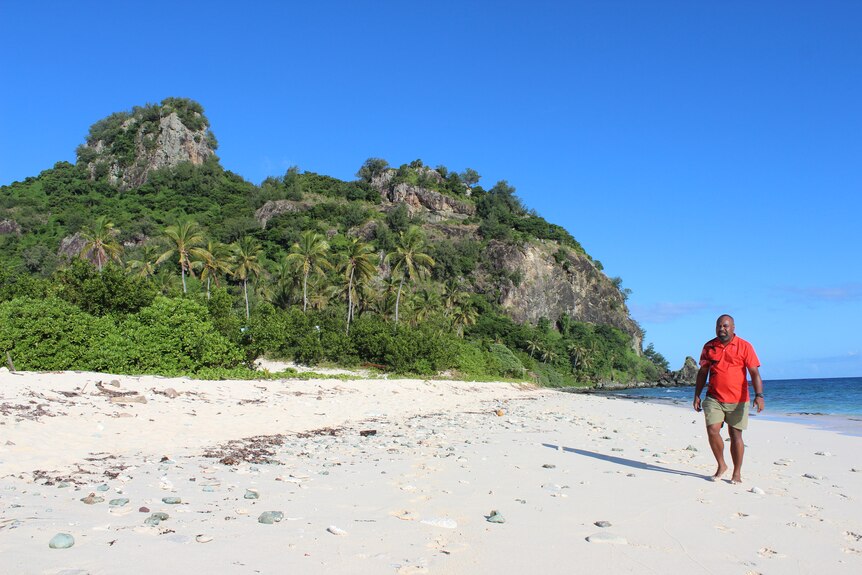 Fijian man in red shirt walks along empty white sandy beach surrounded by palm trees and green shrub. 