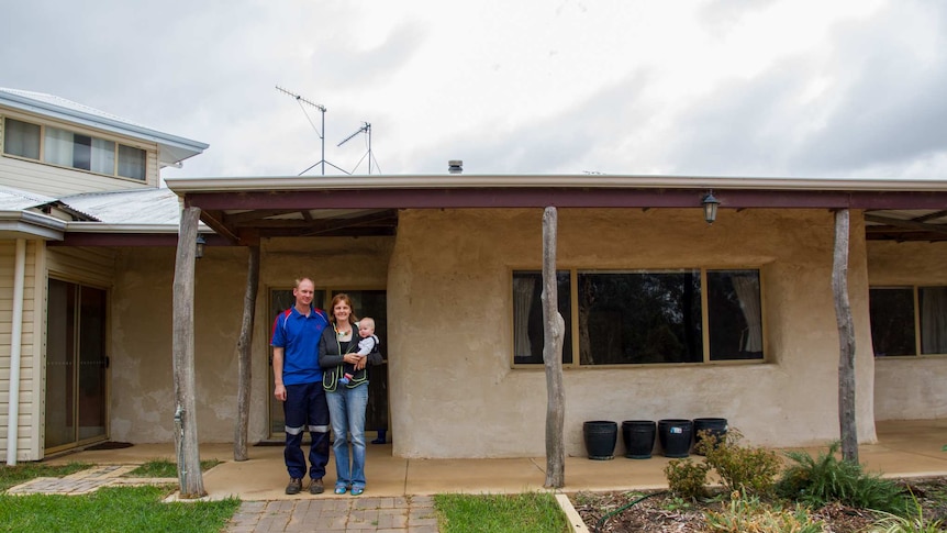 A man and a woman holding a baby stand in front of a house.