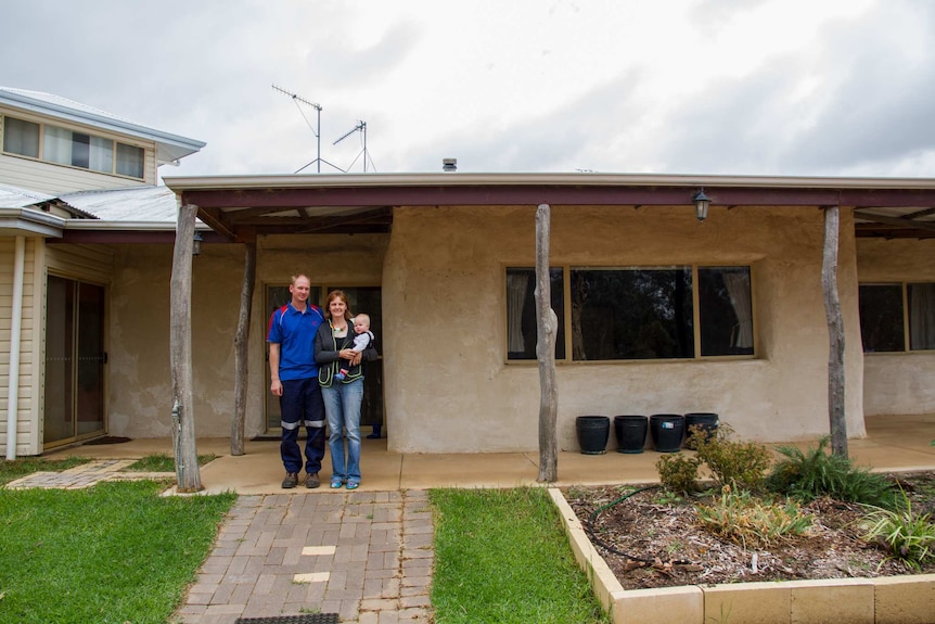 A man and a woman holding a baby stand in front of a house.