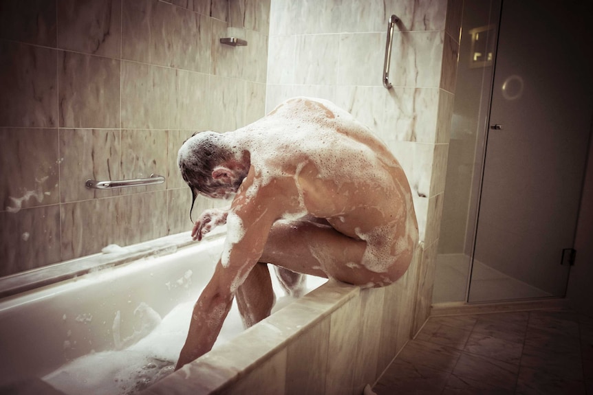 A naked man covered in soap and bubbles, sits in a bath