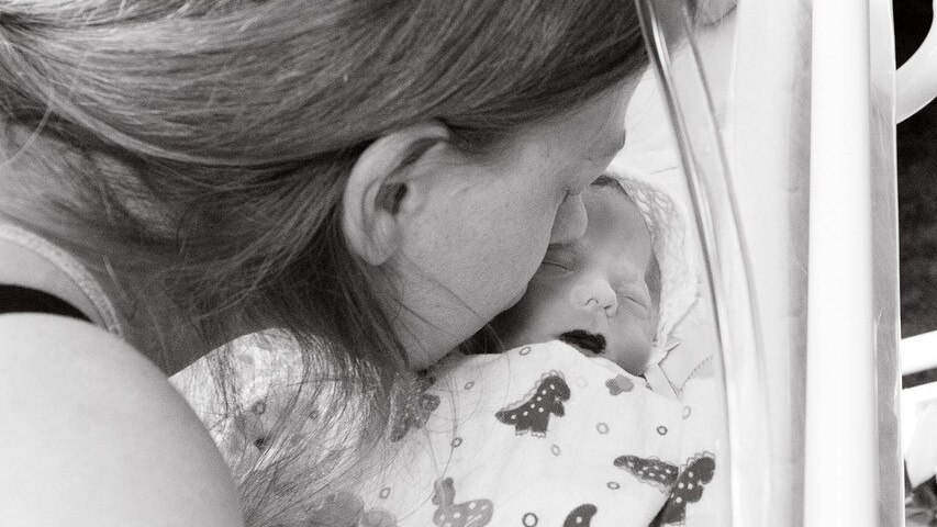 A black and white photo of a mother leaning down to kiss her stillborn baby in a cot.