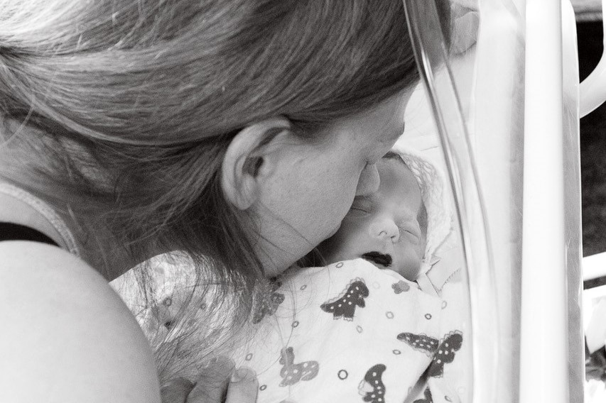 A black and white photo of a mother leaning down to kiss her stillborn baby in a cot.