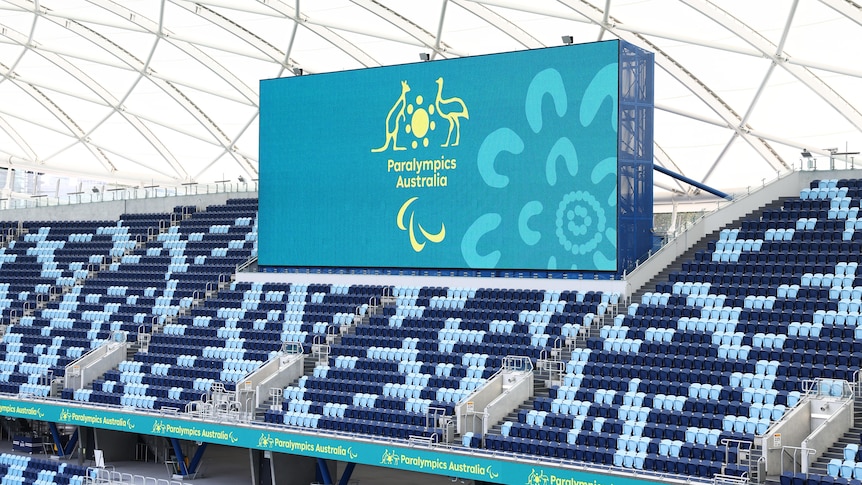 A big screen in an empty stadium shows a gold logo on an aqua background,  saying 'Paralympics Australia'.