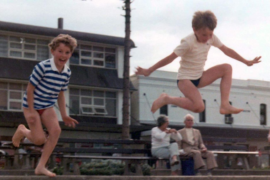 A 1980s photograph of two boys in t-shirts and speedos laughing while jumping high in the air