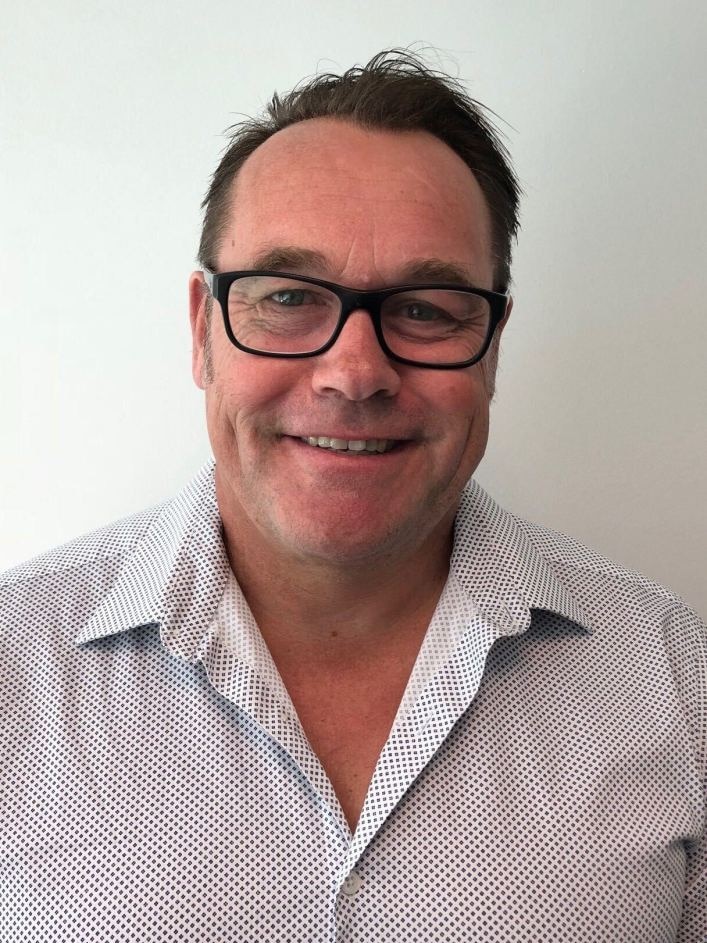 A head shot of a smiling middle-aged man, receding hairline, patterned white shirt, black-rimmed glasses.