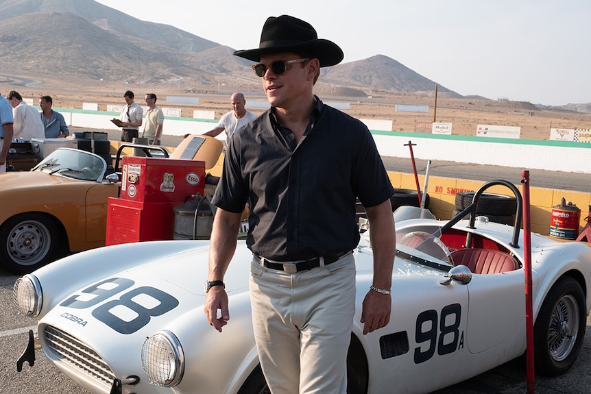 Matt Damon wears black hat and sunglasses and stands in front of vintage racing car and race track near desert landscape.