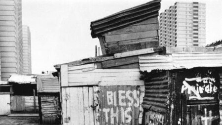 Black and white photos of shacks bunched together with buildings in the background
